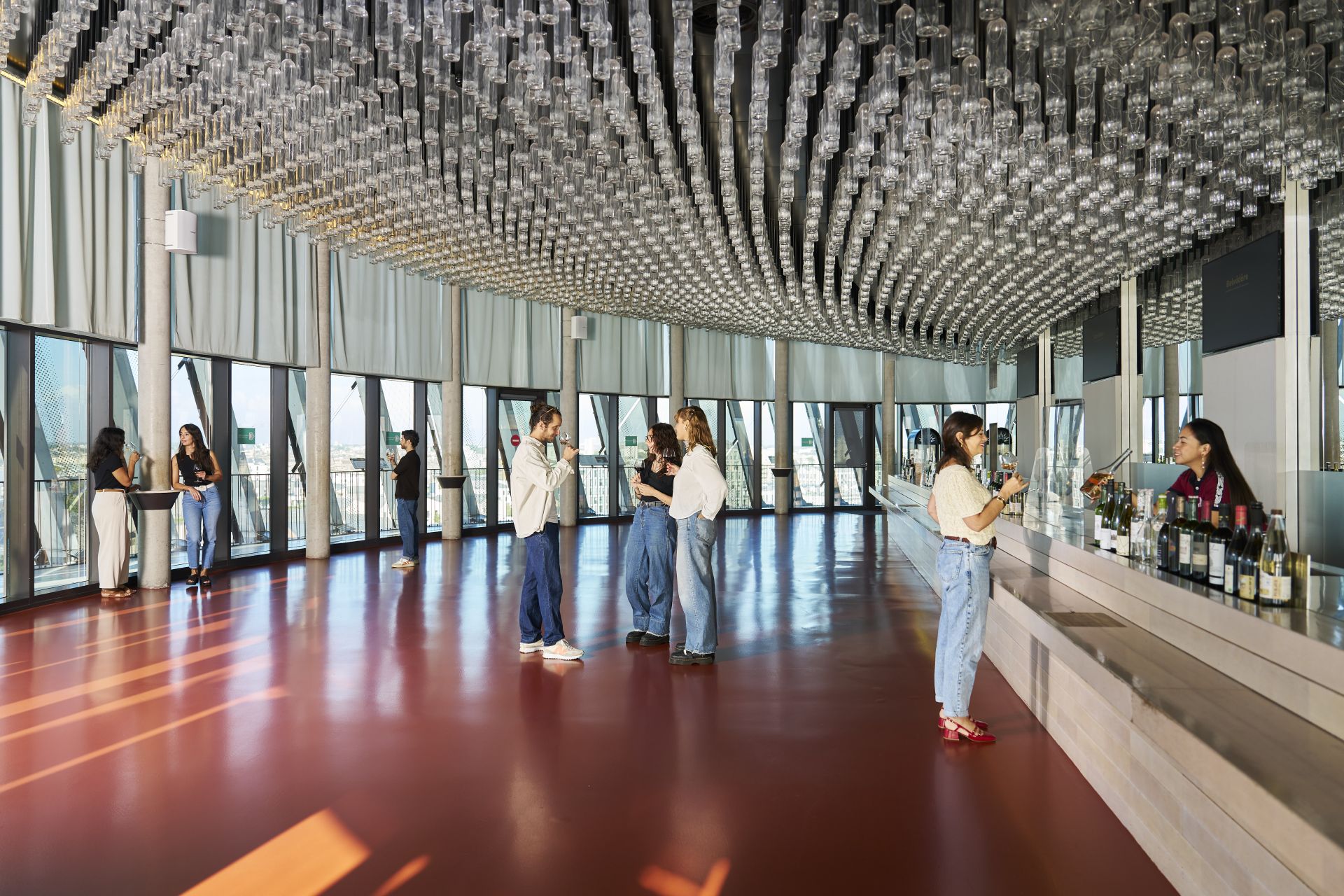 Interior moderno de la Cité du Vin en Bordeaux con visitantes explorando un amplio espacio luminoso, suelo rojo brillante, techo con instalación de botellas de vidrio y grandes ventanales con vistas panorámicas.