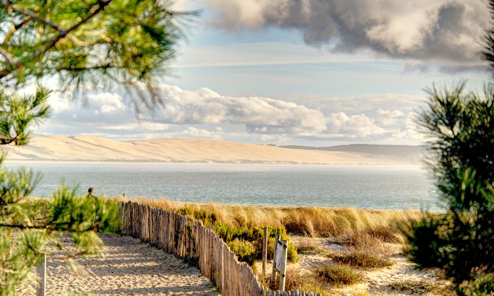 Vista de una playa con dunas de arena, vegetación costera y una valla de madera, con el océano y cielo nublado de fondo