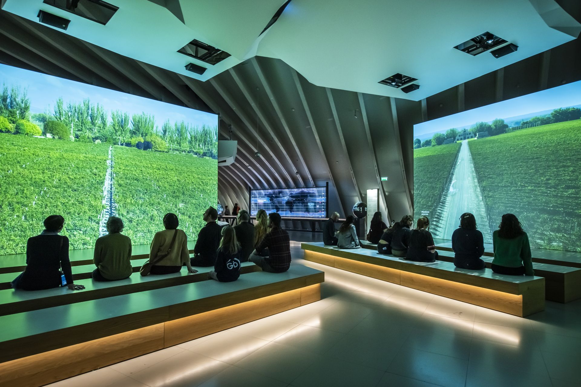 Interior moderno de la Cité du Vin en Burdeos con visitantes sentados frente a pantallas grandes que muestran viñedos y campos de cultivo, bajo un techo con estructura arquitectónica ondulante.