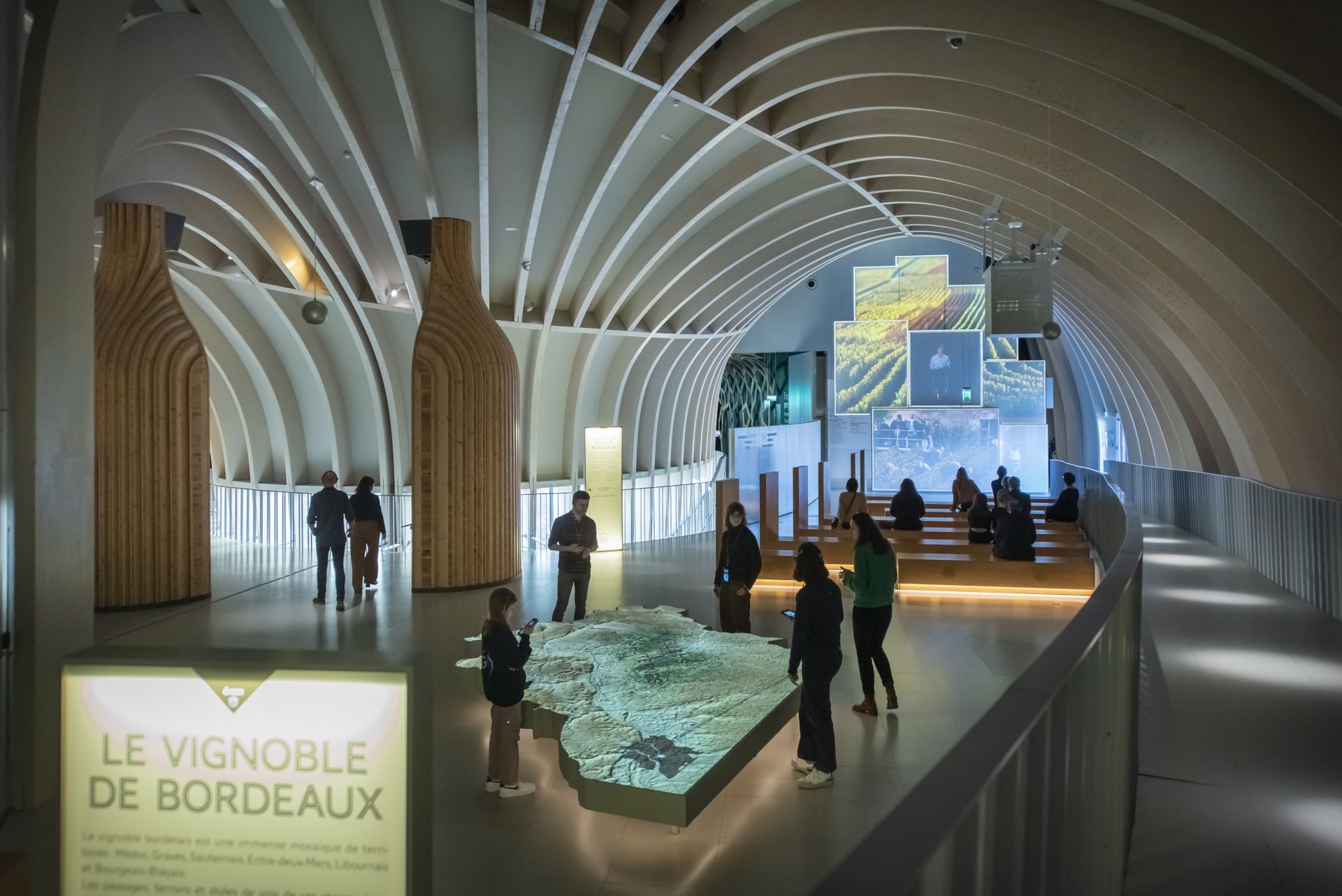 Interior moderno de la Cité du Vin en Burdeos con arquitectura de arcos blancos, visitantes observando una maqueta iluminada del viñedo de Burdeos, paneles informativos y videos sobre viticultura en las paredes