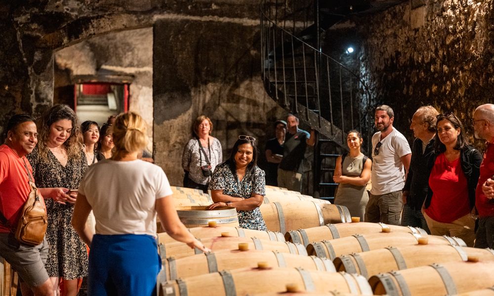 Grupo de turistas visitando una bodega de vinos en Burdeos, observando barriles de roble durante una cata nocturna en las antiguas catacumbas subterráneas.