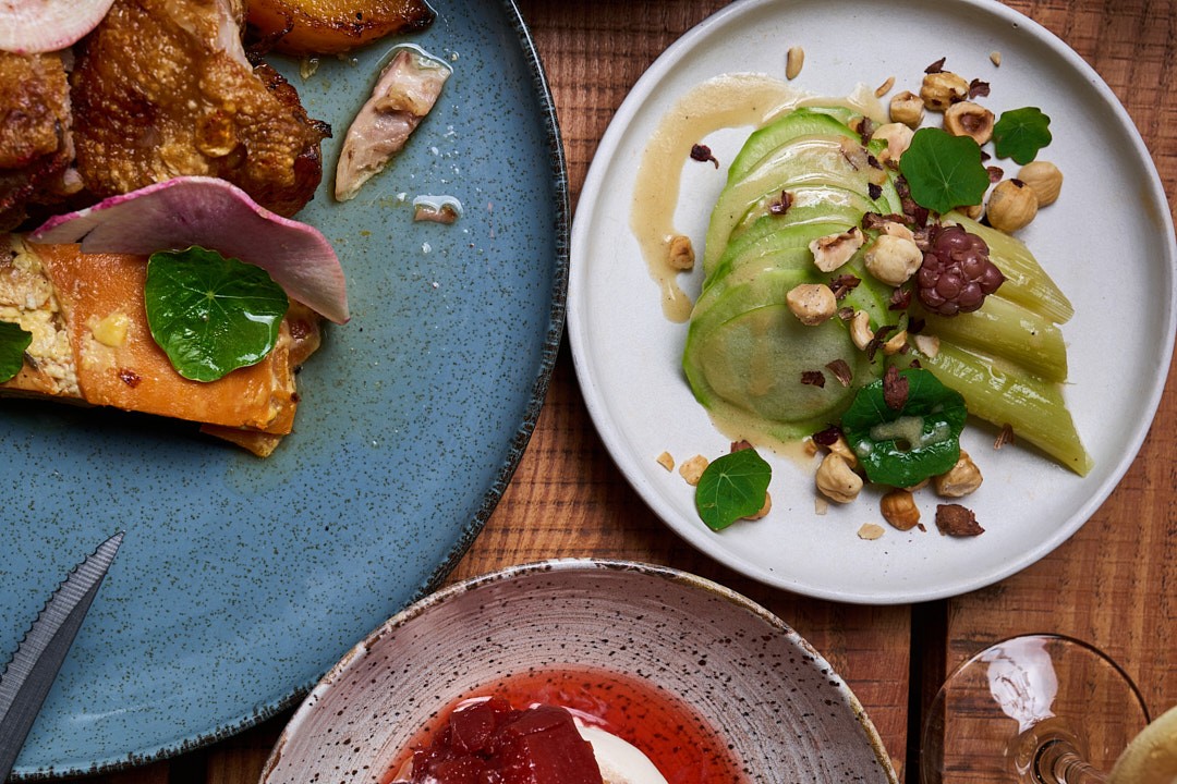 Tres platos de alta cocina presentados en una mesa de madera: a la izquierda, un plato azul con salmón y embutido; en el centro, un plato blanco con aguacate, manzana verde y frutos secos; abajo, un plato texturizado con salsa roja y crema. Decorados con 