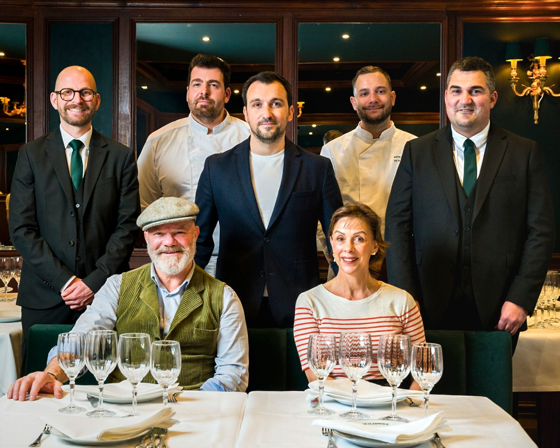 Grupo de siete personas posando en un restaurante elegante de Bordeaux, con dos personas sentadas a una mesa con copas de vino y cinco personas de pie detrás, ante ventanales que muestran la ciudad iluminada por la noche.
