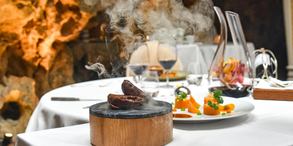 Presentación culinaria gourmet con humo, plato de carne sobre base de madera y piedra, acompañamientos de verduras en plato blanco, copas de vino tinto de fondo, en restaurante de lujo