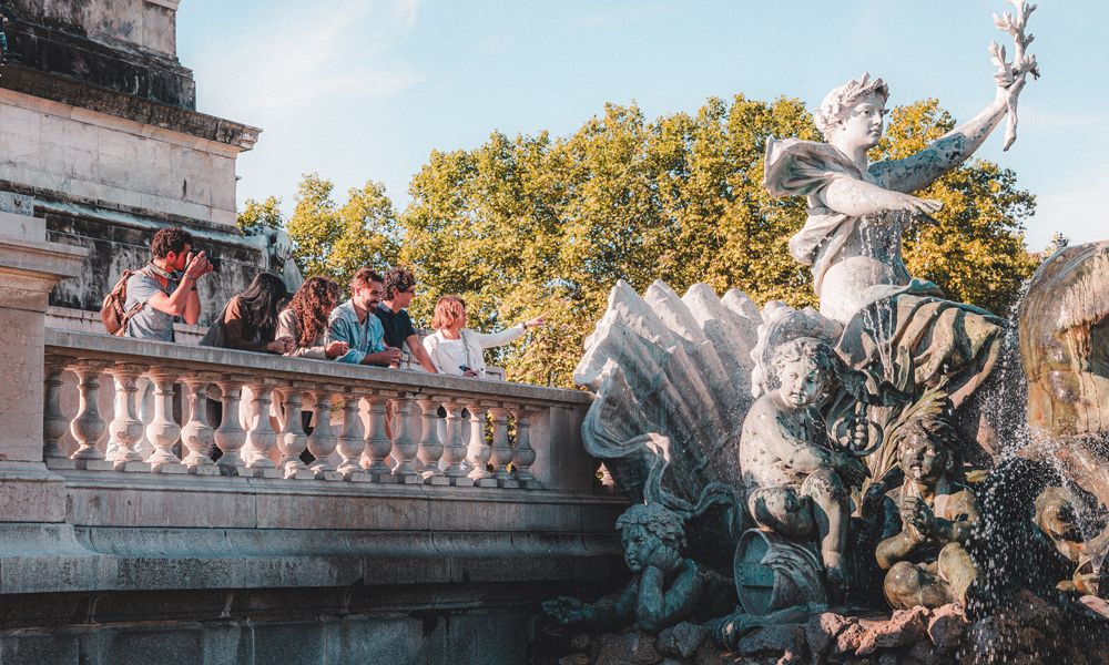 Fuente monumental en una plaza de Burdeos con estatuas de bronce verdoso, turistas observando desde una terraza con barandilla, y árboles frondosos de color dorado al fondo bajo un cielo despejado.