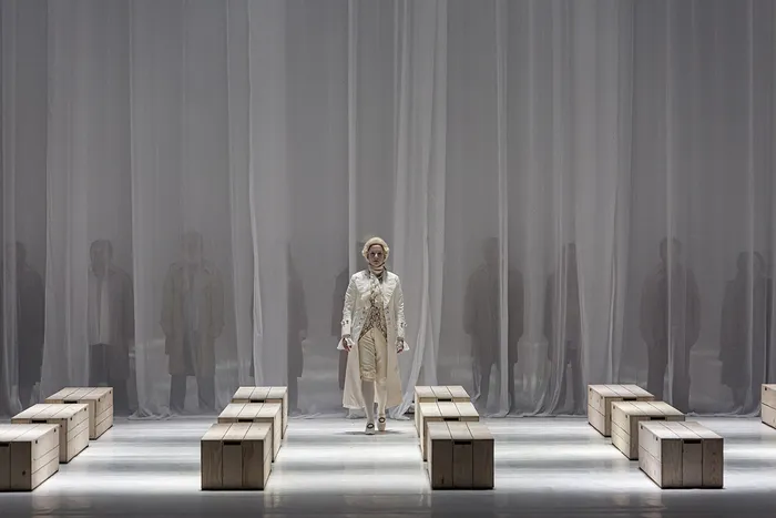 Un solista en traje de color claro y sombrero se encuentra en el centro del escenario en una escena minimalista con bloques de madera y telón gris estriado, rodeado de figuras borrosas, en una producción del Réquiem de Mozart en Burdeos.