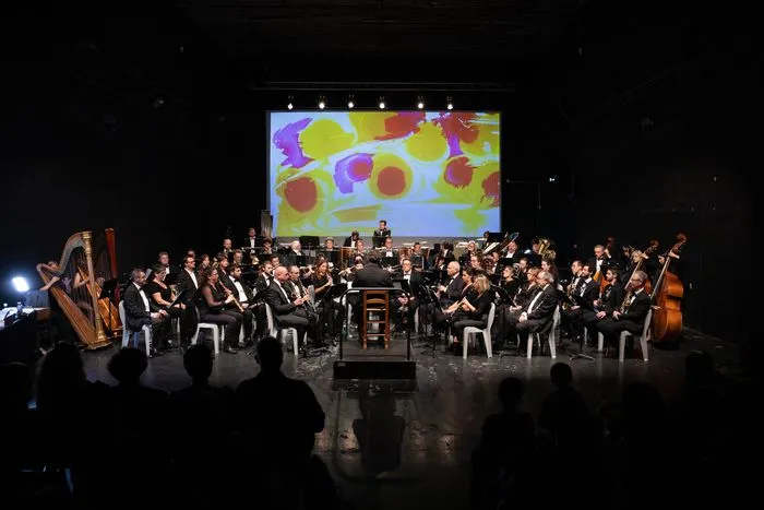 La Orquesta de Viento de Burdeos en concierto en escena, con músicos en traje formal tocando diversos instrumentos, una pantalla de proyección mostrando formas coloridas al fondo, y un director de orquesta dirigiendo.