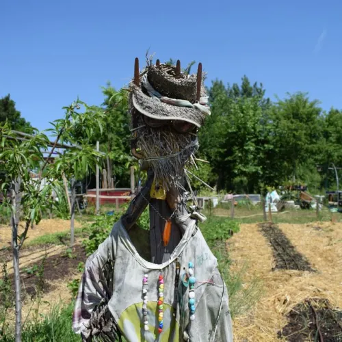 Espantapájaros artístico con camisa blanca y cuentas de colores, con cabeza de paja coronada de ramas, de pie en un jardín comunitario de Burdeos.