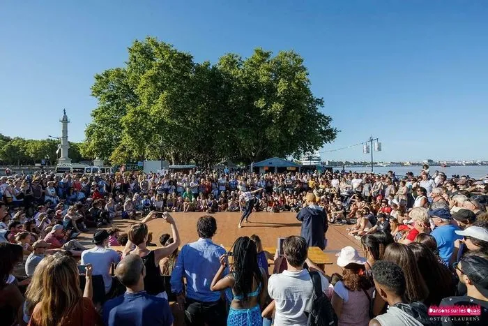 Gran multitud de personas reunidas en los muelles de Burdeos para un evento festivo de danza al aire libre, con árboles, un monumento y el puerto de fondo bajo un cielo azul despejado.
