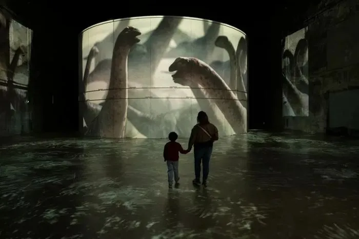 Dos visitantes, un niño y un adulto, tomados de la mano en una sala inundada de los Bassins des Lumières en Burdeos, frente a proyecciones gigantes de dinosaurios en las paredes y el techo de la exposición inmersiva.