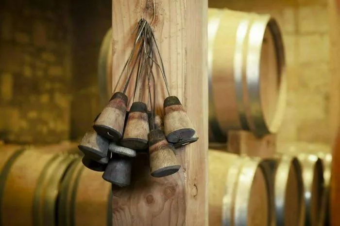 Campanillas de vendimia colgadas de una viga de madera en una bodega con barriles de vino de fondo, durante las Jornadas de Puertas Abiertas de los castillos de Castillon Côtes de Bordeaux