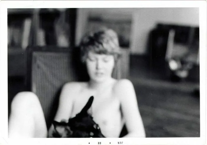 Fotografía en blanco y negro de una mujer sentada con una prenda sin mangas en un interior desenfocado. Imagen de archivo digitalizada fechada en 1969.