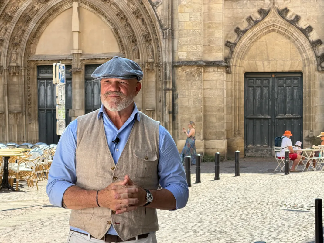 El chef Philippe Etchebest posando frente a una catedral gótica en una plaza adoquinada de Burdeos, vistiendo camisa azul, chaleco beige y gorra plana.