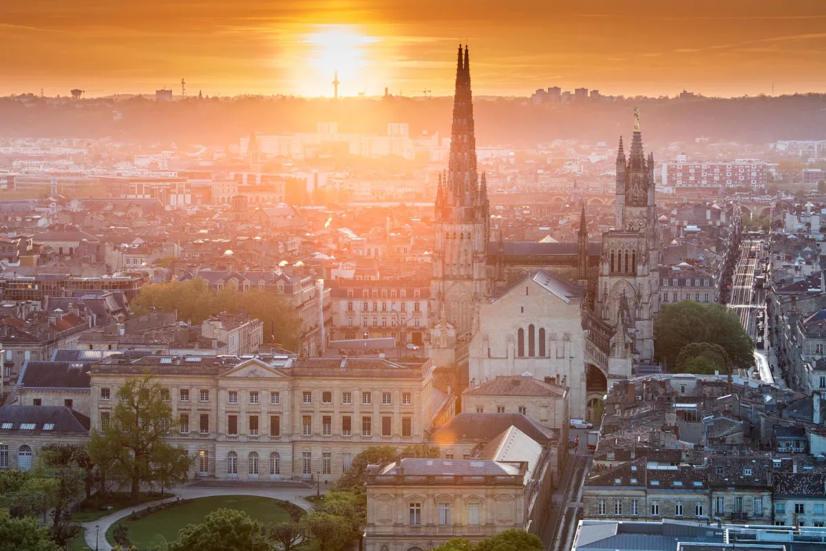 Vista aérea de Burdeos al atardecer con la Basílica de San Miguel y sus dos agujas góticas en primer plano, dominando la arquitectura clásica de la ciudad y sus barrios históricos.
