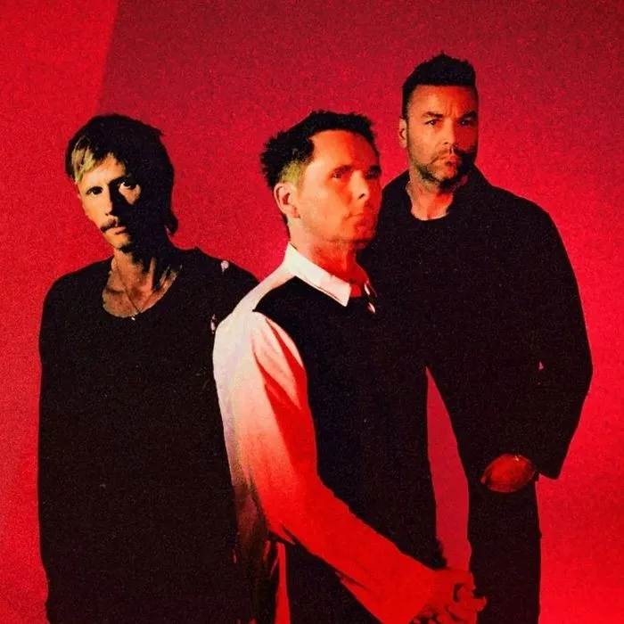 Retrato de tres músicos de la banda Muse en ropa oscura sobre un fondo rojo vibrante para la gira en Burdeos