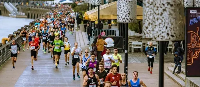 Cientos de corredores participan en una carrera de 10 km en los muelles de Burdeos, luciendo dorsales de colores variados, con multitudes y edificios históricos al fondo.
