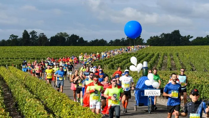 Grupo de corredores del Maratón de los Châteaux du Médoc atravesando un viñedo de Burdeos con un globo azul gigante y carteles de apoyo.