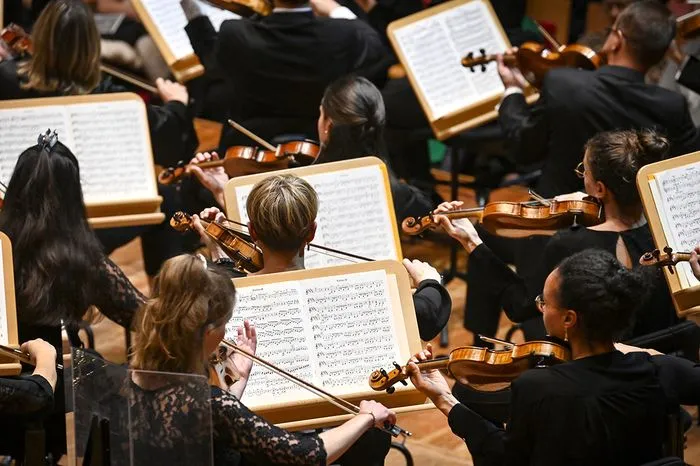 Vista aérea de una orquesta sinfónica durante un ensayo o concierto, con músicos vestidos de negro sosteniendo violines y partituras en atriles.