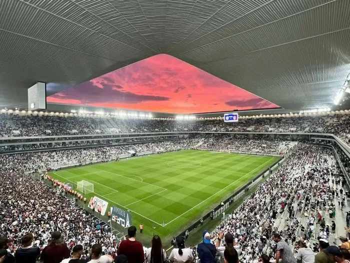 Vista panorámica de un estadio de fútbol lleno de espectadores durante un partido entre Francia y Turquía en Burdeos, con una puesta de sol roja espectacular visible sobre el techo del estadio.