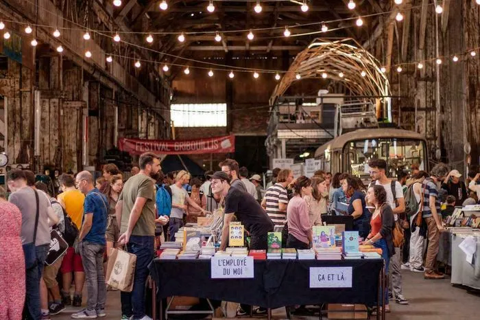 Multitud de visitantes en el Festival Gribouillis de Burdeos, explorando puestos de libros y productos artesanales en un almacén industrial iluminado con luces decorativas.