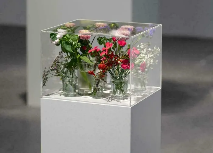 Instalación artística compuesta por una vitrina transparente que contiene varios jarrones de cristal llenos de flores frescas coloridas (rosas, claveles rojos y blancos, respiros de bebé) sobre un pedestal blanco, exposición Pollen en CAPC Burdeos.