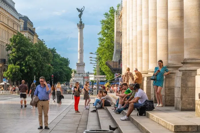 Plaza de la Bolsa de Burdeos con sus imponentes columnas neoclásicas, visitantes descansando en las escaleras y el Monumento a los Girondinos al fondo bajo cielo azul.
