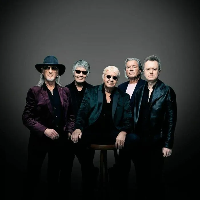 Retrato grupal de cinco músicos de Deep Purple con atuendos negros de escena y accesorios, posando sobre fondo gris.