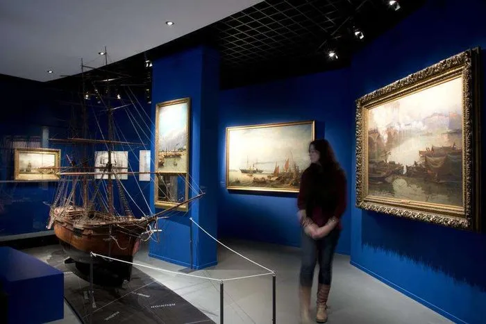 Galería de museo con paredes azul oscuro, maqueta de barco antiguo, cuadros de marinas enmarcados en dorado, y visitante observando las obras de arte
