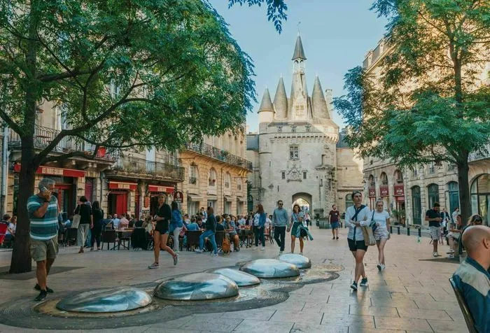 La Puerta Cailhau, monumento histórico de Burdeos, con sus torres puntiagudas características, rodeada de una plaza pública animada con visitantes y árboles verdes.