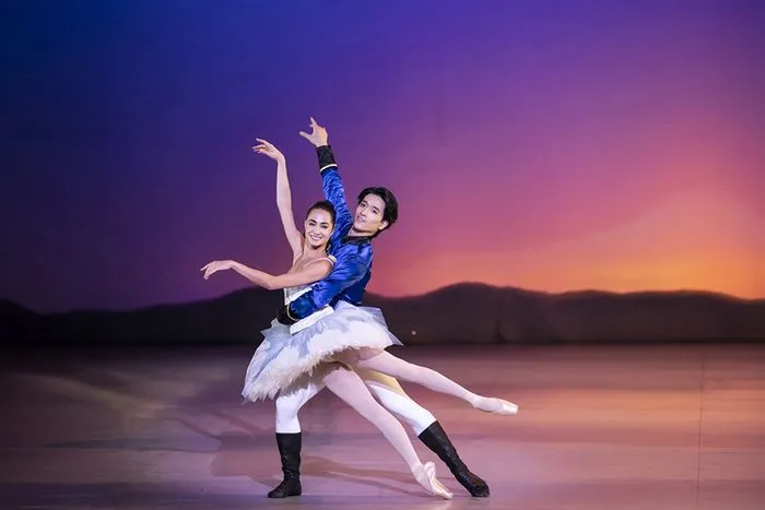 Dos bailarines de ballet en traje de príncipe y bailarina ejecutan un pas de dos en el escenario con un fondo de atardecer.