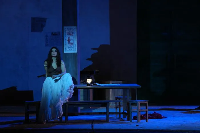 Escena de la ópera Carmen en el Teatro Nacional de Burdeos: una mujer con traje tradicional sentada en una mesa iluminada bajo luces azules, con un escenario minimalista y escaleras de fondo.
