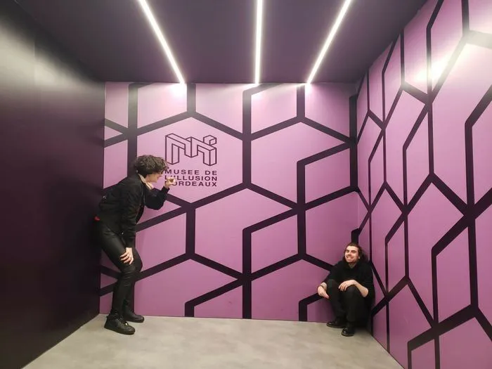 Dos visitantes en una sala del Museo de la Ilusión de Burdeos, rodeados de paredes rosas con patrones geométricos negros e iluminados por luces neón blancas en el techo.