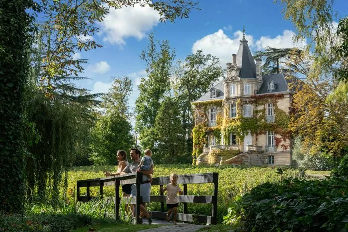 Familia observando un castillo vinícola desde una terraza de madera en los viñedos de Graves, cerca de Burdeos, con vistas a una hermosa mansión de estilo Belle Époque rodeada de vegetación.