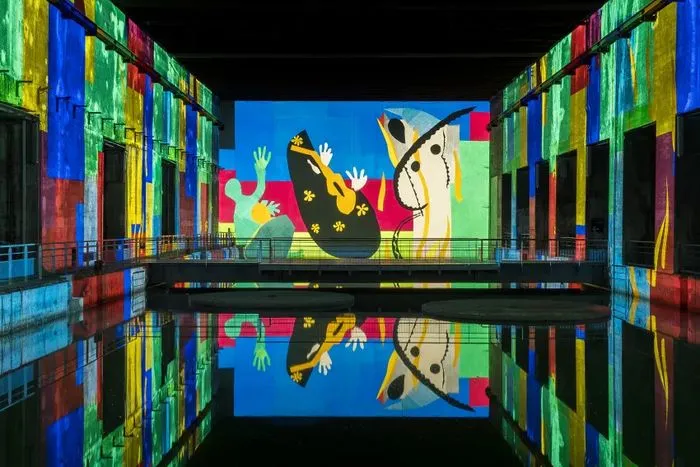 Instalación inmersiva en los Bassins des Lumières de Burdeos que presenta una obra colorida inspirada en Matisse, con figuras abstractas en tonos vibrantes (azul, rojo, amarillo, verde) proyectadas en las paredes y reflejadas en el agua.