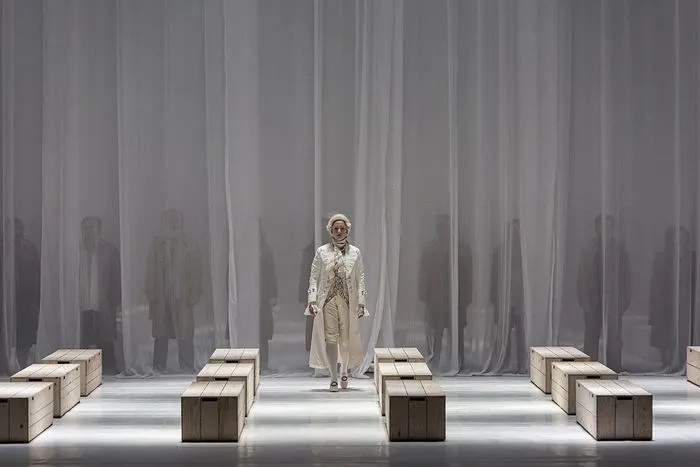 Un solista en traje de color claro y sombrero se encuentra en el centro del escenario en una escena minimalista con bloques de madera y telón gris estriado, rodeado de figuras borrosas, en una producción del Réquiem de Mozart en Burdeos.