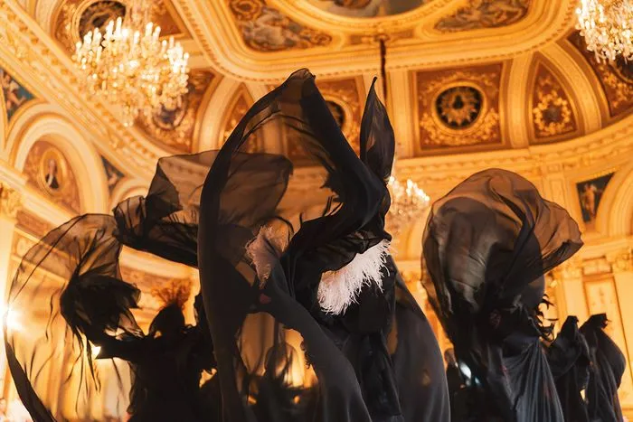 Siluetas de personajes vestidos de negro con velos y sombreros puntiagudos bajo un techo dorado ornamentado con lámpara de cristal en el Baile de las Brujas de Burdeos.