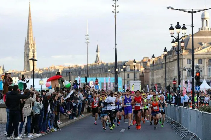 Corredores participando en la media maratón de Burdeos, corriendo en una carretera cerrada con espectadores a lo largo del recorrido y la arquitectura histórica de Burdeos de fondo.