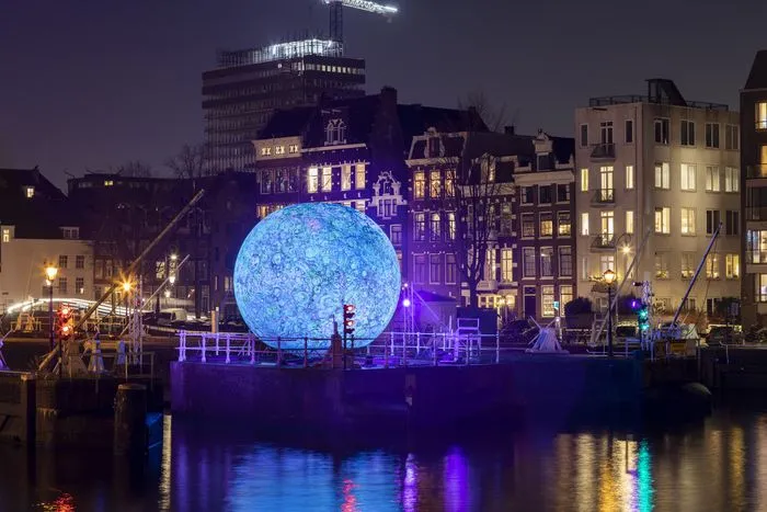 Instalación artística de una esfera azul gigante iluminada en los muelles de Burdeos por la noche, rodeada de edificios históricos iluminados en púrpura, reflejada en el agua del río.
