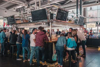 Vista interior de las Halles de Bacalan en Burdeos con una multitud de visitantes alrededor de una barra de vinos. Se ven estantes de vasos suspendidos y pizarras de menú sobre el mostrador en este mercado cubierto moderno.