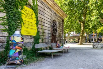 Patio empedrado con edificios históricos de piedra cubiertos de hiedra, cartel amarillo « Conviértete en arte vivo », escultura colorida, bancos públicos y persona sentada, árboles de fondo