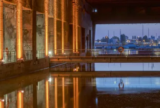 Vista nocturna de la Base Submarina de Burdeos con columnas iluminadas en naranja reflejadas en agua tranquila, con vistas al puerto y el río Garona al atardecer.