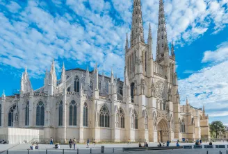 La Catedral de Saint-André de Burdeos, edificio gótico blanco con dos torres principales, fachada ornamentada con vitrales y rosetones, rodeada de visitantes en un patio empedrado bajo un cielo azul nublado.