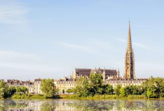 Vista panorámica de la Basílica de San Miguel de Burdeos con su campanario gótico dominando el paisaje urbano, rodeada de edificios de estilo Haussmann y reflejada en las aguas del río Garona.