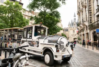 Un tren turístico eléctrico blanco y negro circulando por una calle adoquinada de Burdeos, con edificios de estilo Haussmann y un castillo al fondo.