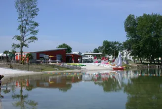 Vista de Playa Bègles en Burdeos con un edificio rojo junto al agua, velas de colores, árboles y una persona en primer plano