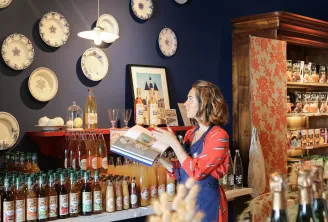 Una vendedora presenta productos en la Echoppe de la Lune en Burdeos, rodeada de botellas de vino y licores, con platos decorativos azules y blancos colgados en la pared.