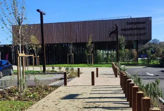 Entrada del complejo acuático Elodie Lorandi en Burdeos con edificio moderno de madera y acero, paseo pavimentado bordeado de bolardos, árboles y espacios verdes