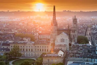 Vista aérea de Burdeos al atardecer con la Basílica de San Miguel y sus dos agujas góticas en primer plano, dominando la arquitectura clásica de la ciudad y sus barrios históricos.