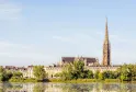 Vista panorámica de la Basílica de San Miguel de Burdeos con su campanario gótico dominando el paisaje urbano, rodeada de edificios de estilo Haussmann y reflejada en las aguas del río Garona.