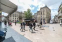 Plaza animada de Burdeos con peatones, ciclistas y tiendas, bordeada de edificios de estilo haussmaniano bajo un cielo nublado.